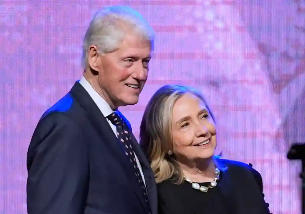 Bill and Hillary Clinton smiling and posing together at an event with Bill Clinton wearing a suit and Hillary Clinton in a dark-colored outfit.