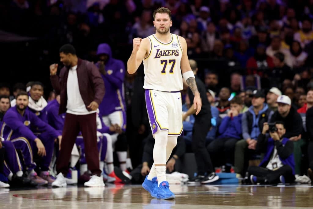 Luka Dončić on the court in a Lakers uniform, fist raised during a game with teammates and fans in the background.