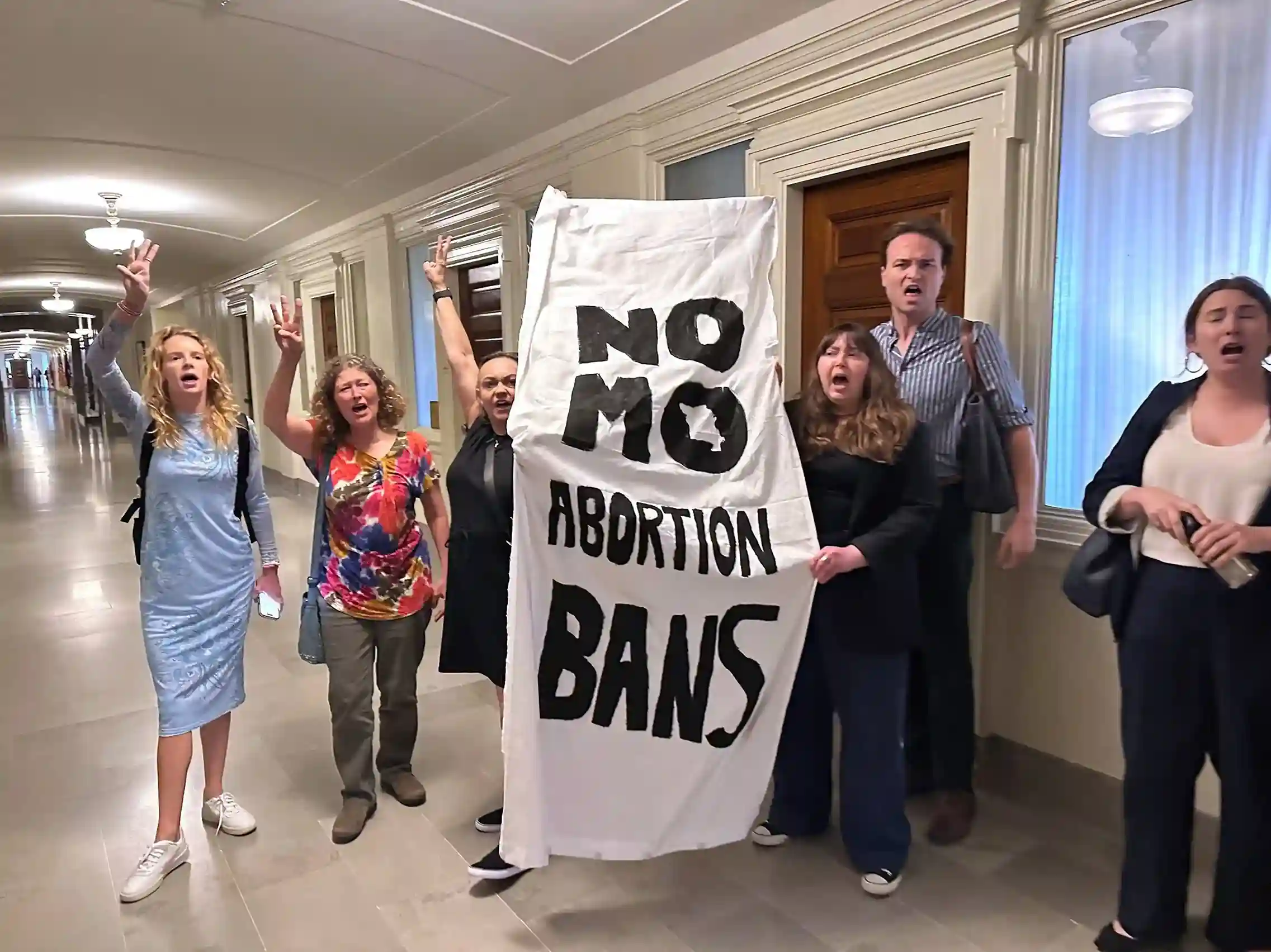 Group of activists holding a banner that reads 'No More Abortion Bans' while protesting inside a government building hallway.
