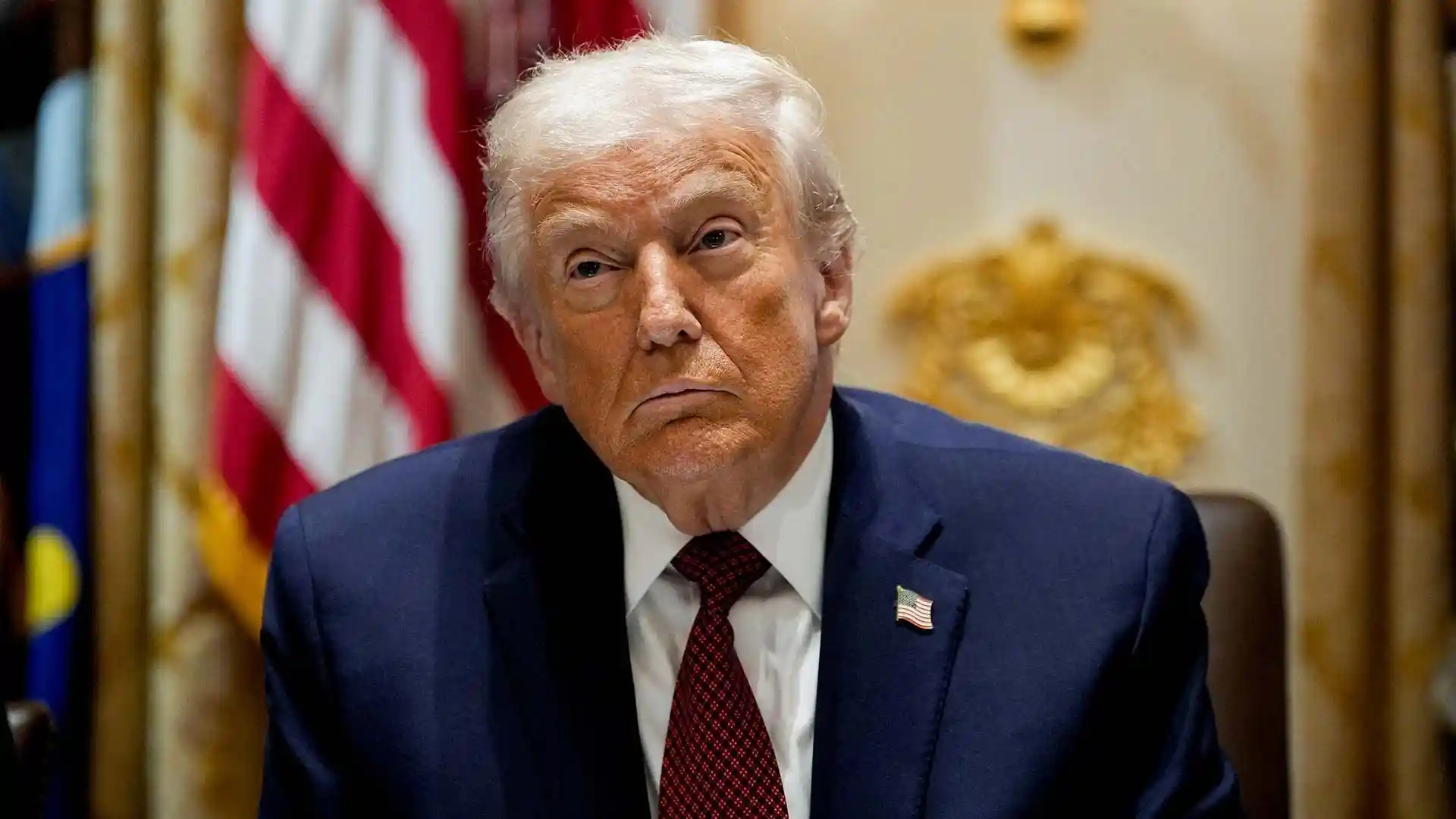 Donald Trump sitting during a formal meeting with the U.S. flag in the background, looking serious