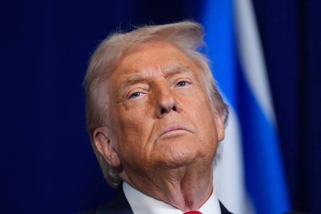 A drone in flight over a coastal area at dusk, with explosions in the distance near a remote dock facility.