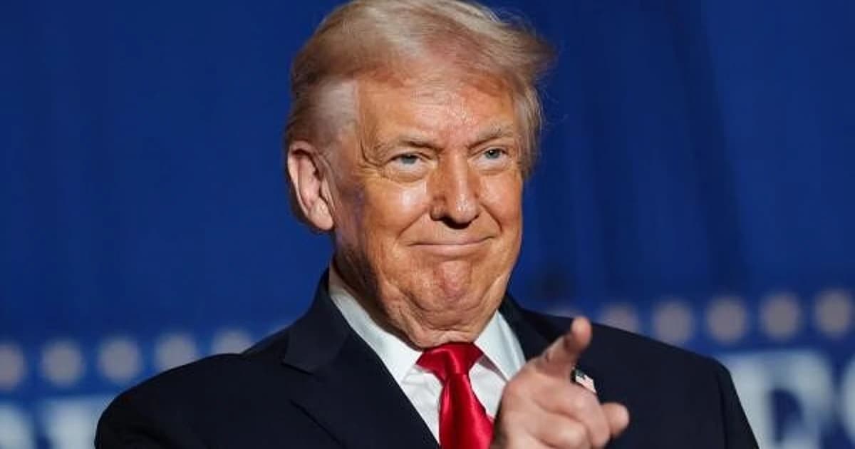 Donald Trump smiling and pointing forward during a public appearance, with a blue backdrop behind him.